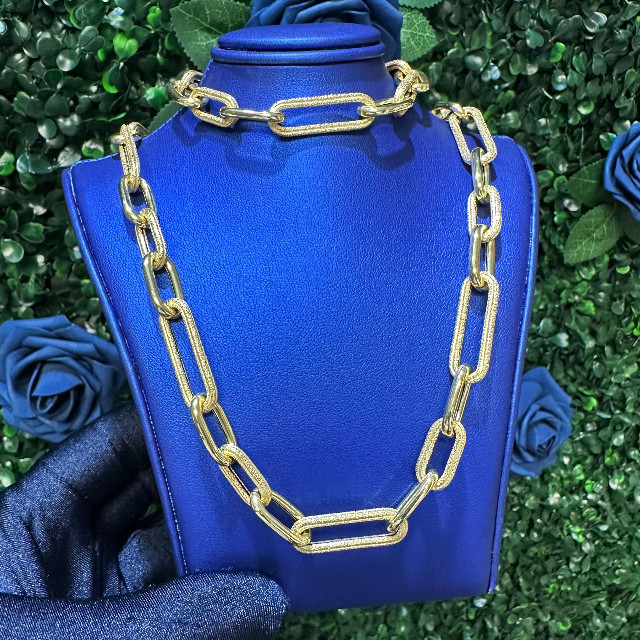 Gold chain necklace and bracelet set on a blue mannequin with green foliage in the background