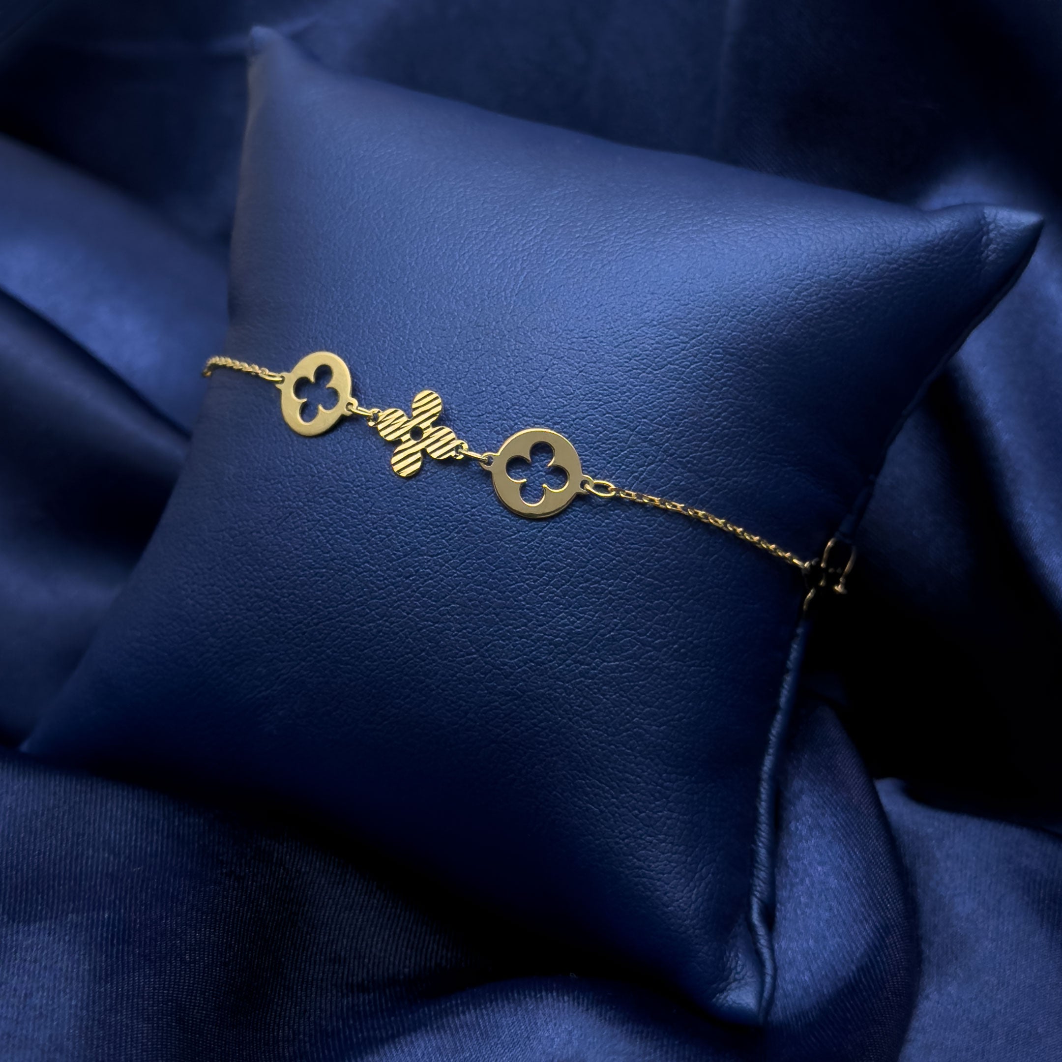 A delicate gold chain bracelet featuring four leaf clover charms with round cutout clover motifs, displayed on a dark blue satin pillow against a matching blue fabric background.