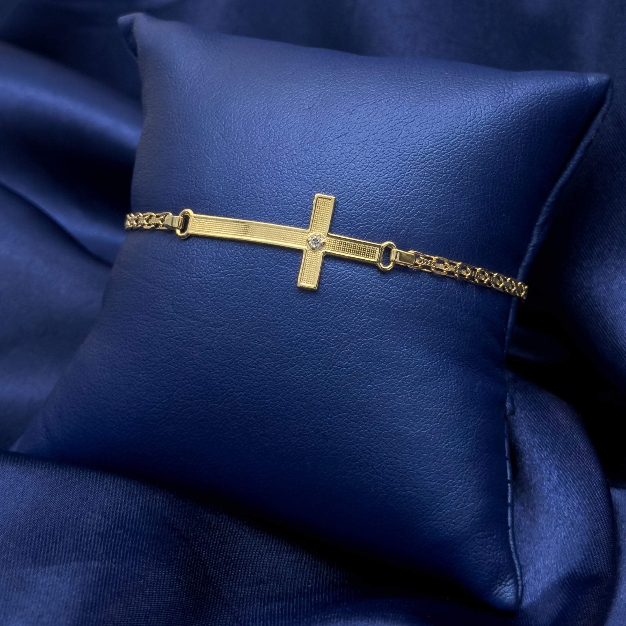 A gold cross bracelet with a small central gemstone, displayed on a textured dark blue satin pillow.