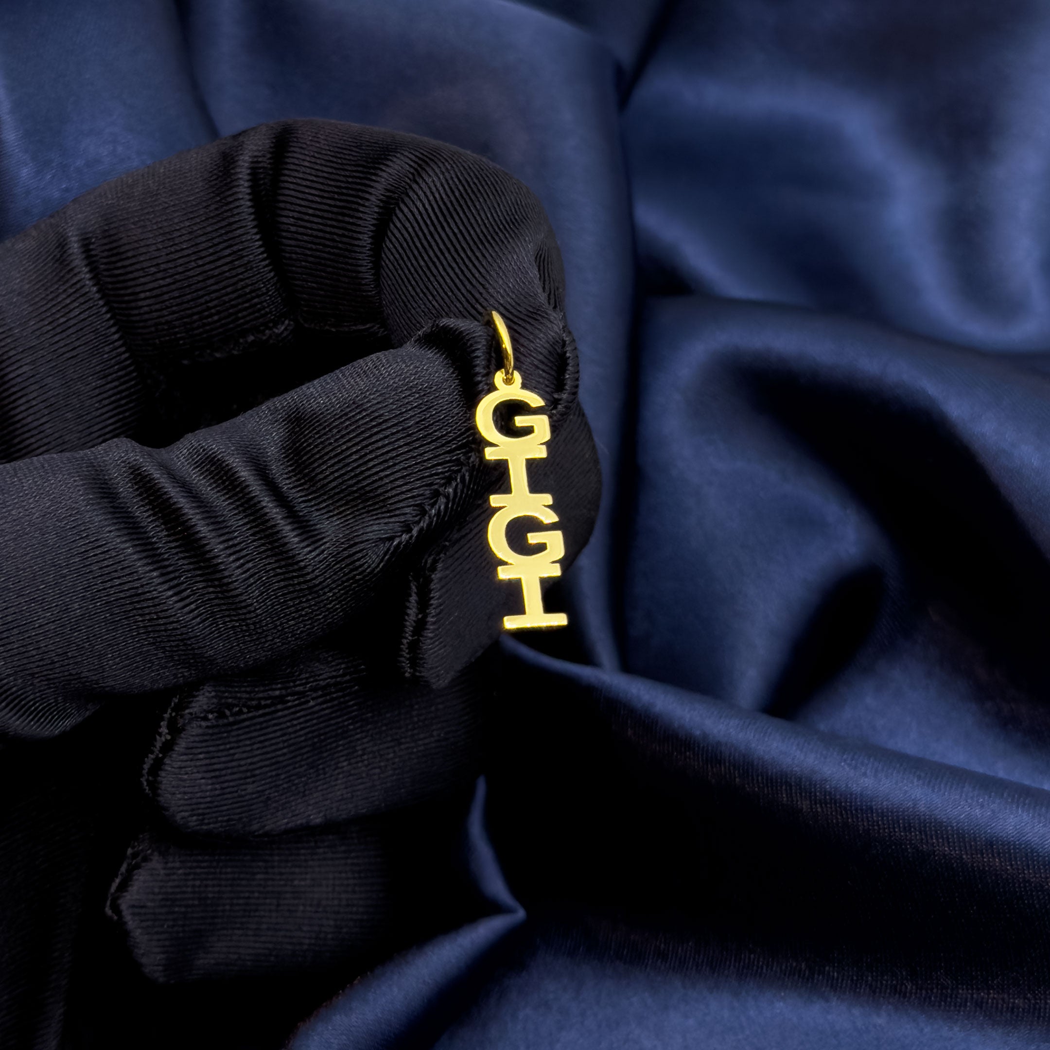 Blue glove holding a gold pendant with 'Gigi' in 14k gold on a blue fabric background
