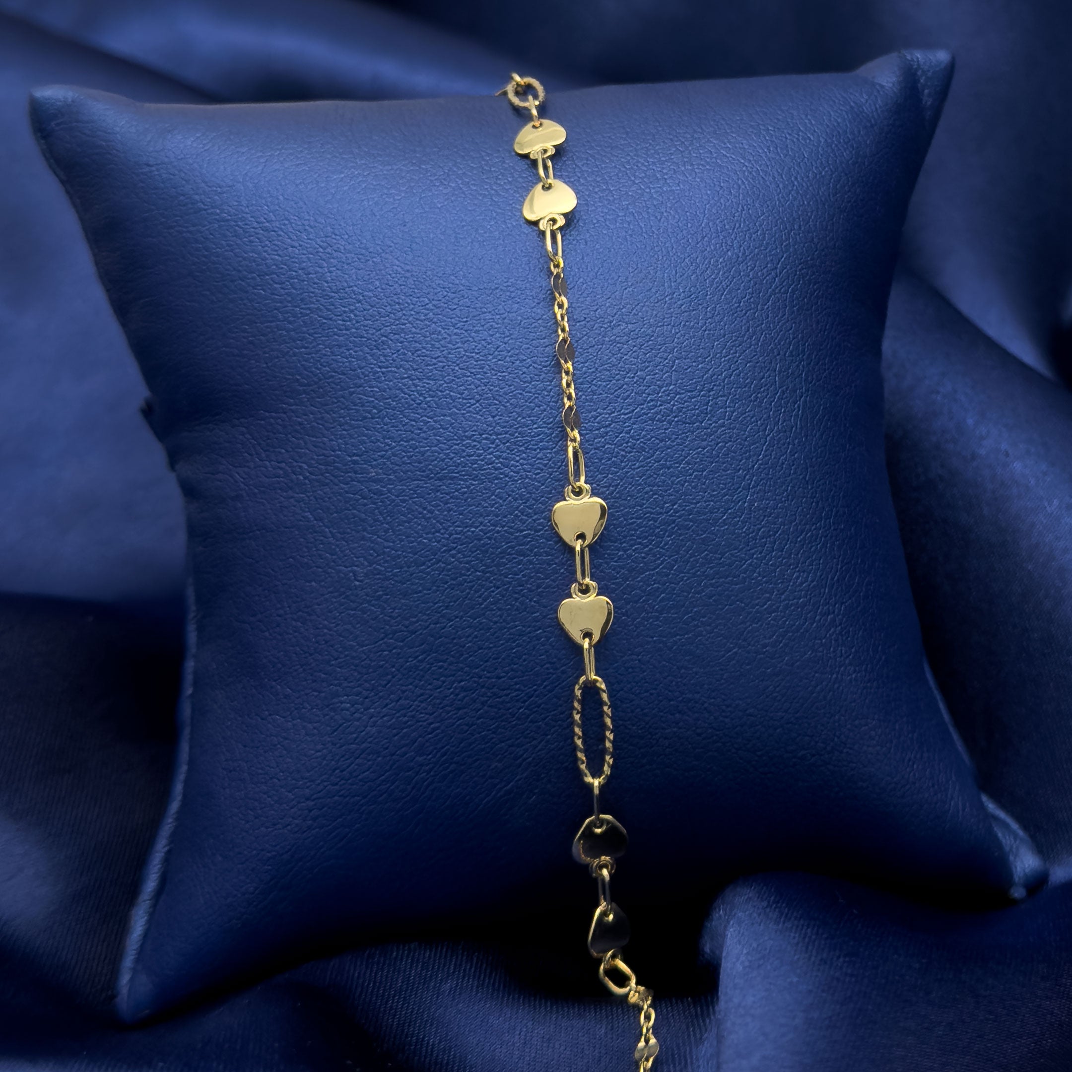A gold heart-link bracelet draped over a dark blue satin pillow on a matching blue fabric background.