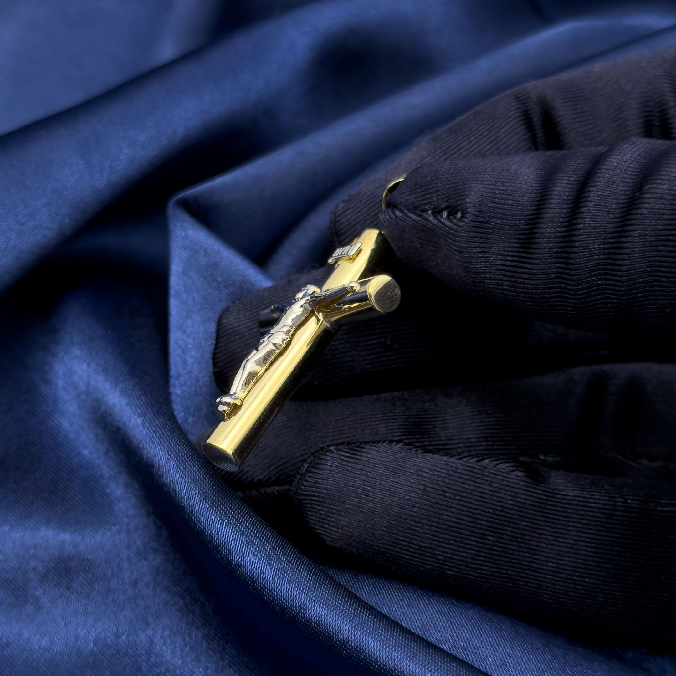 A gold crucifix pendant with a white gold toned figure of Jesus, held by a hand wearing a blue glove over draped dark blue satin fabric. side angle