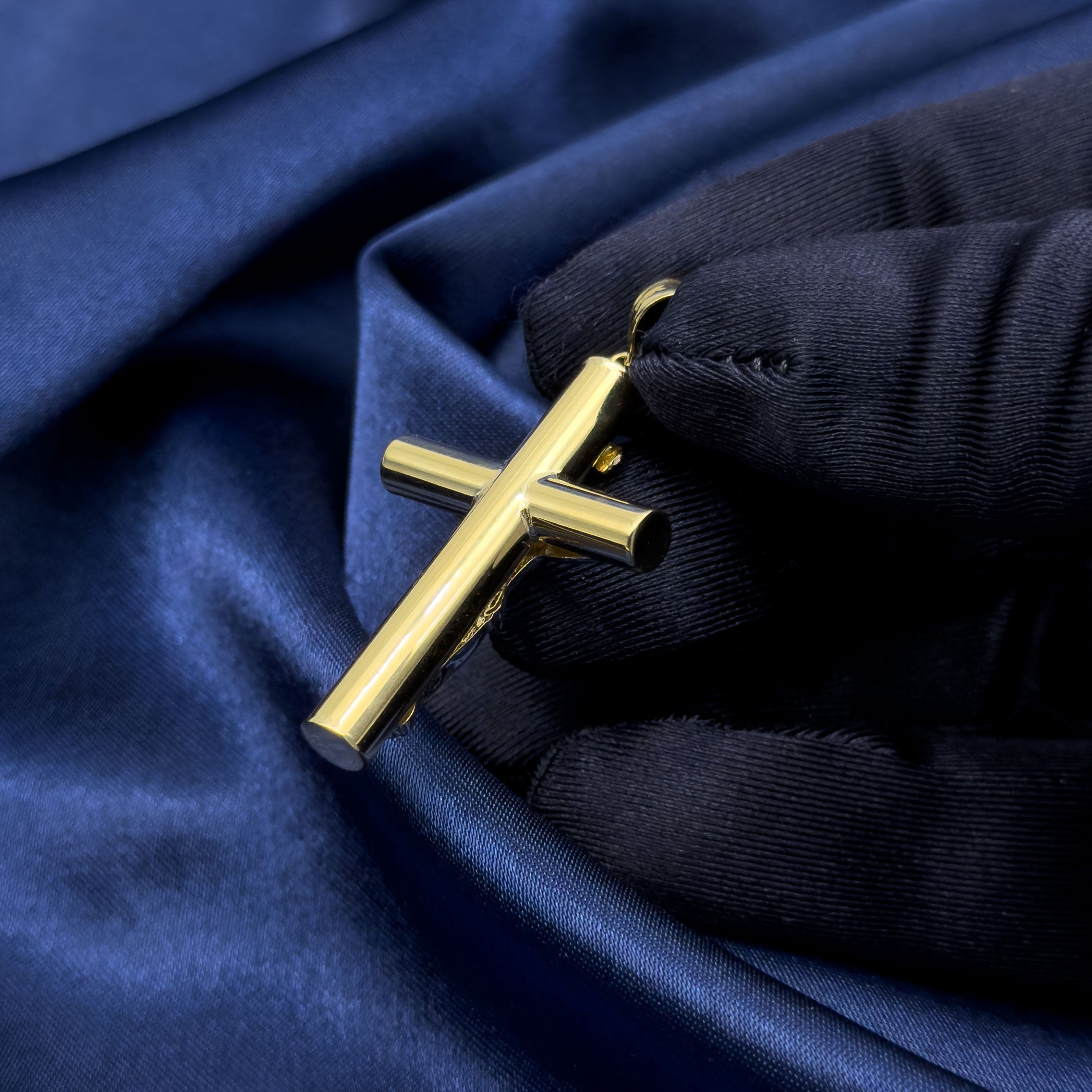A gold crucifix pendant with a white gold toned figure of Jesus, held by a hand wearing a blue glove over draped dark blue satin fabric. back
