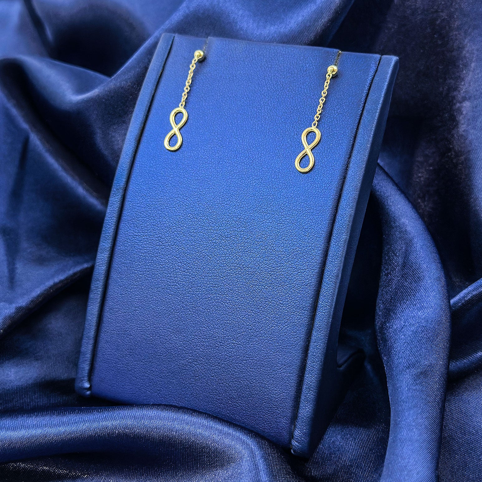 14k yellow gold hanging infinity earrings on blue earring display with a blue background