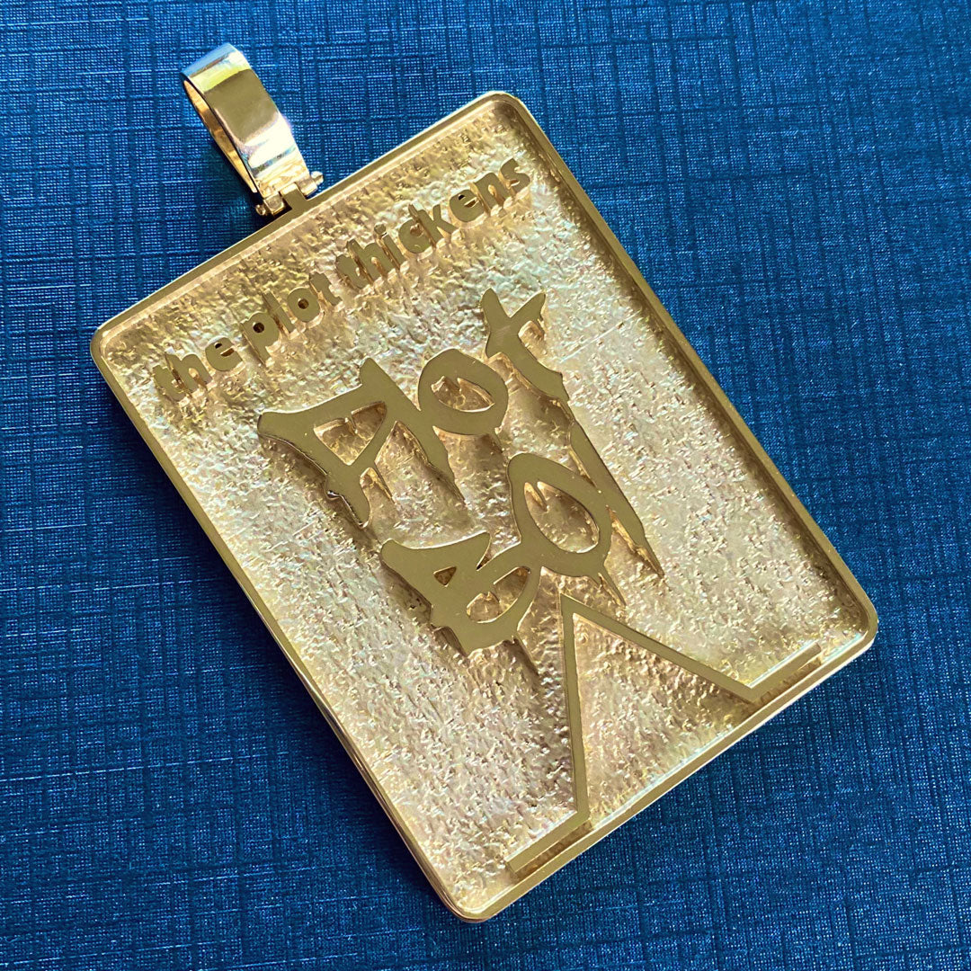 Gold pendant with 'Plot Boi' design on a blue textured background