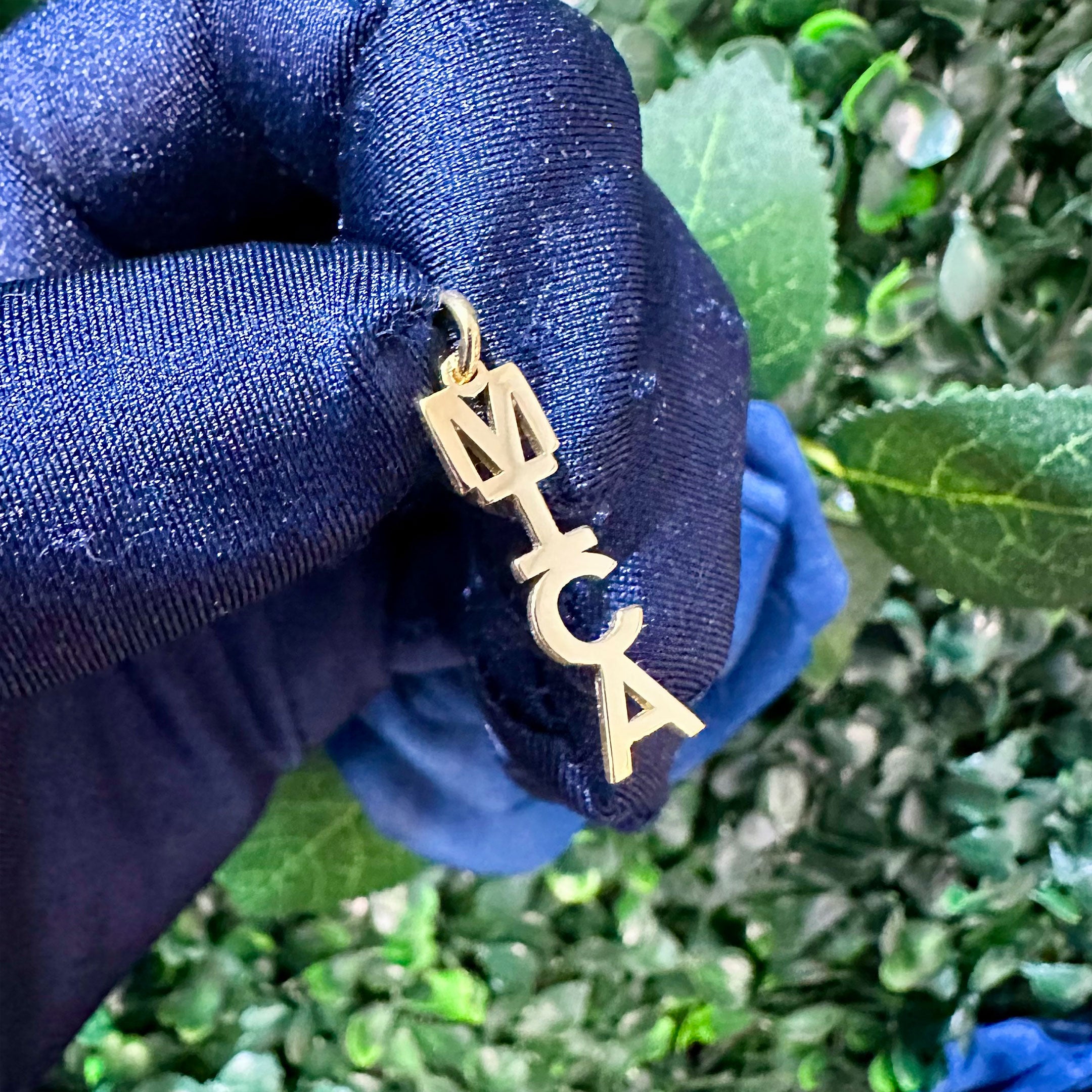 Gold pendant with 'MICA' letters held by a gloved hand against a green leafy background