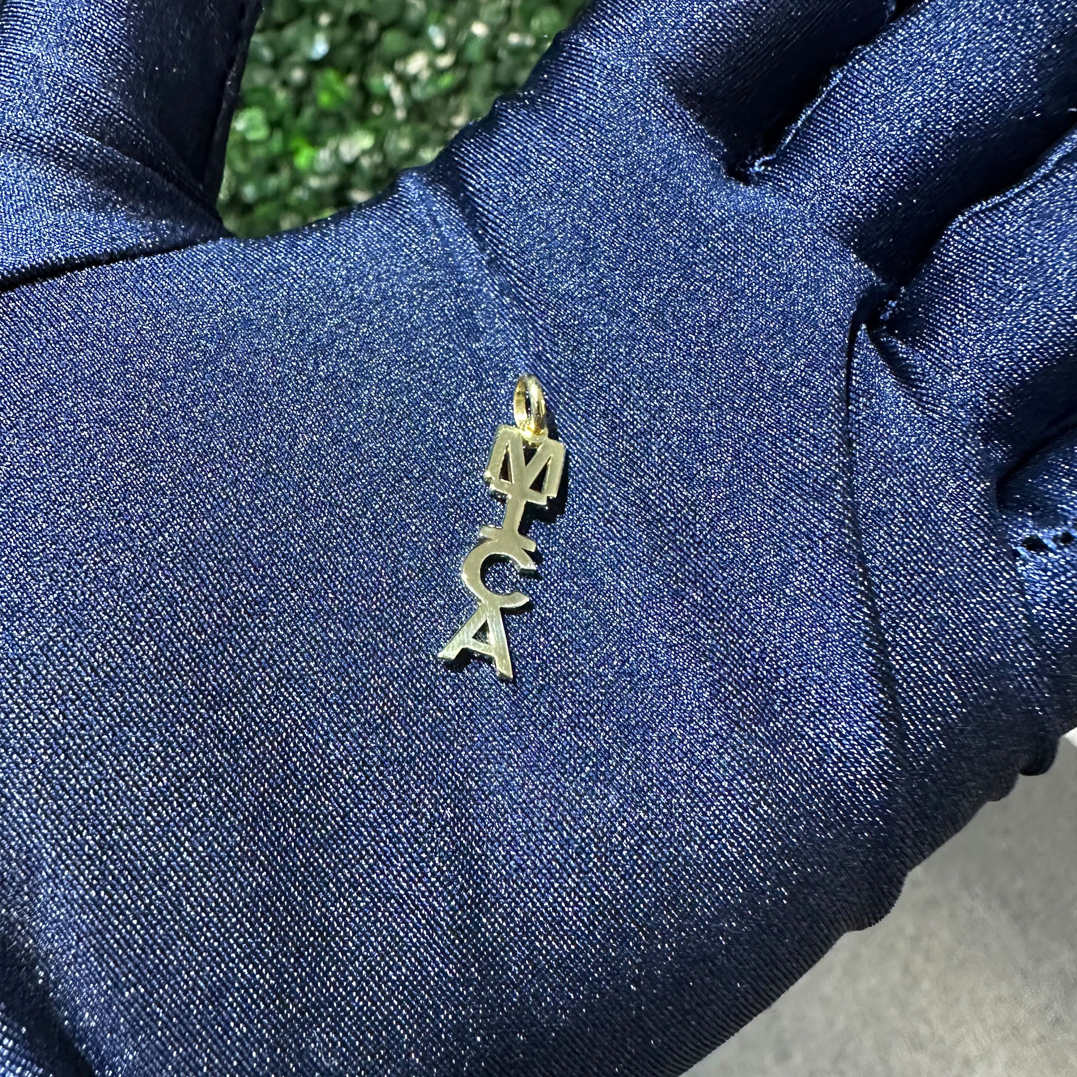 Gold pendant with 'MICA' letters held by a gloved hand in blue glove hand
