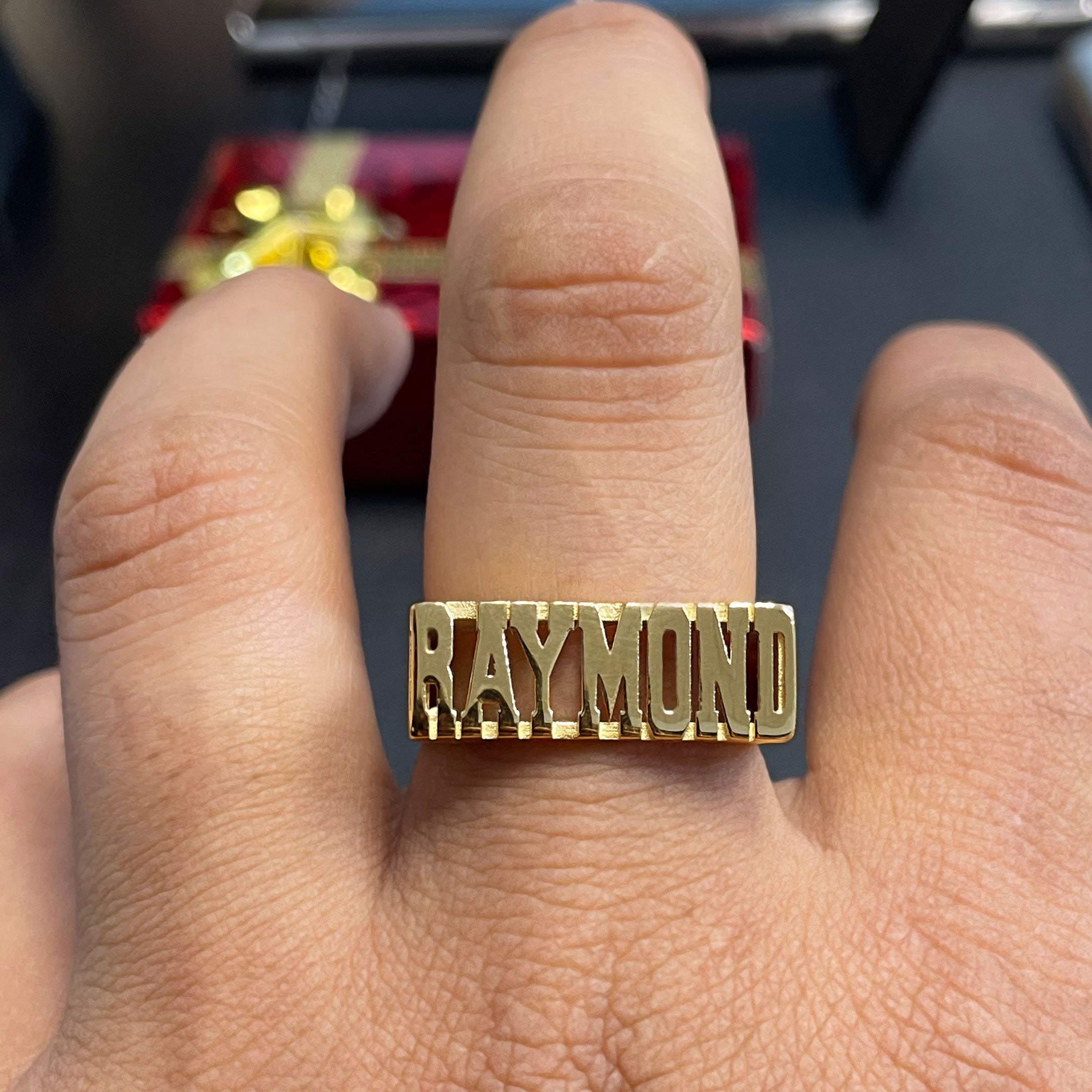 Gold ring with 'RAYMOND' on a finger against a blurred background