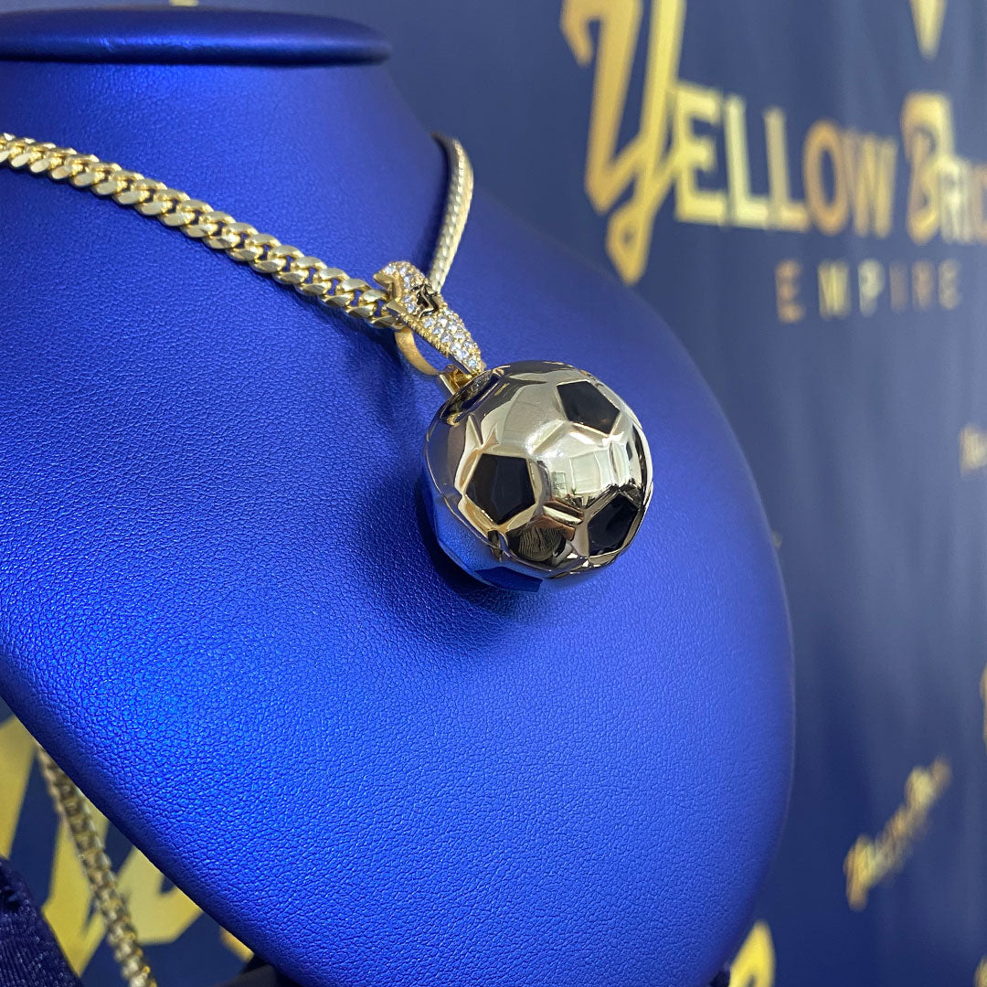 Gold necklace with a soccer ball pendant on a blue surface, branded 'Yellow Brick Empire'.
