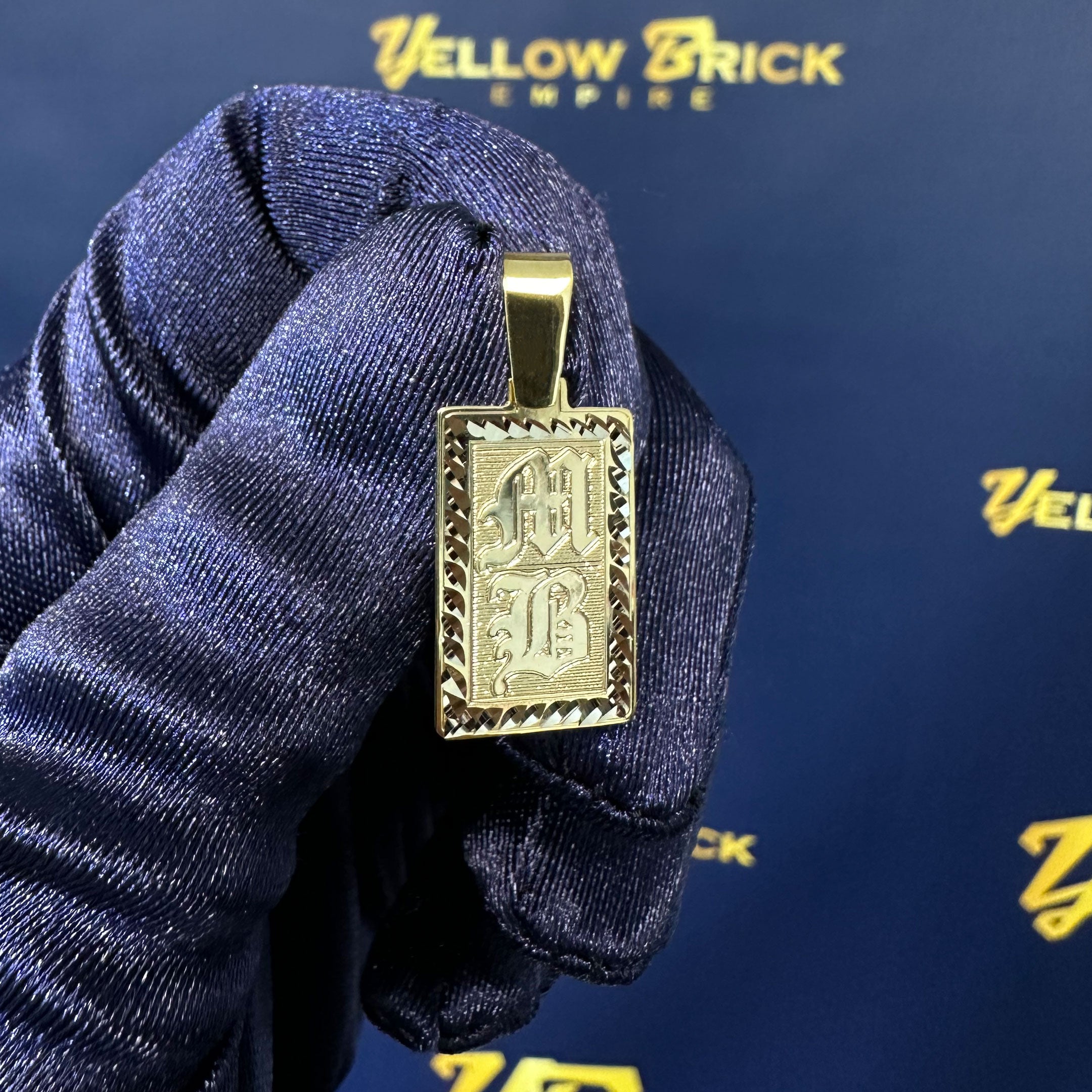 Gold rectangle pendant with diamond cut border and MB engraved in Old English in Hand with Blue Glove