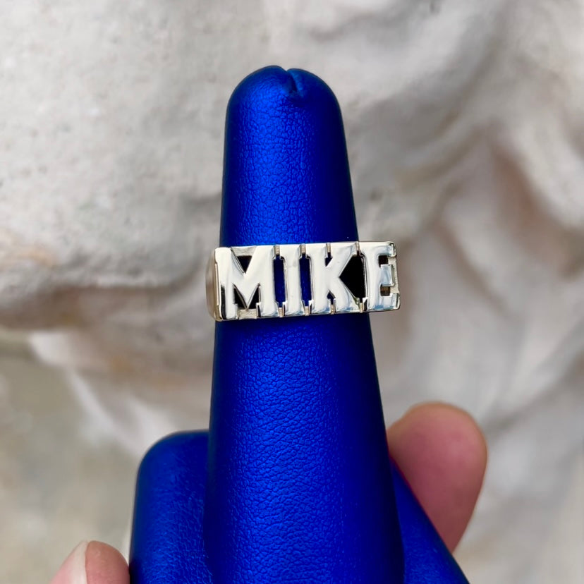 A yellow gold ring with the name 'MIKE' in block letters, displayed on a blue stand.