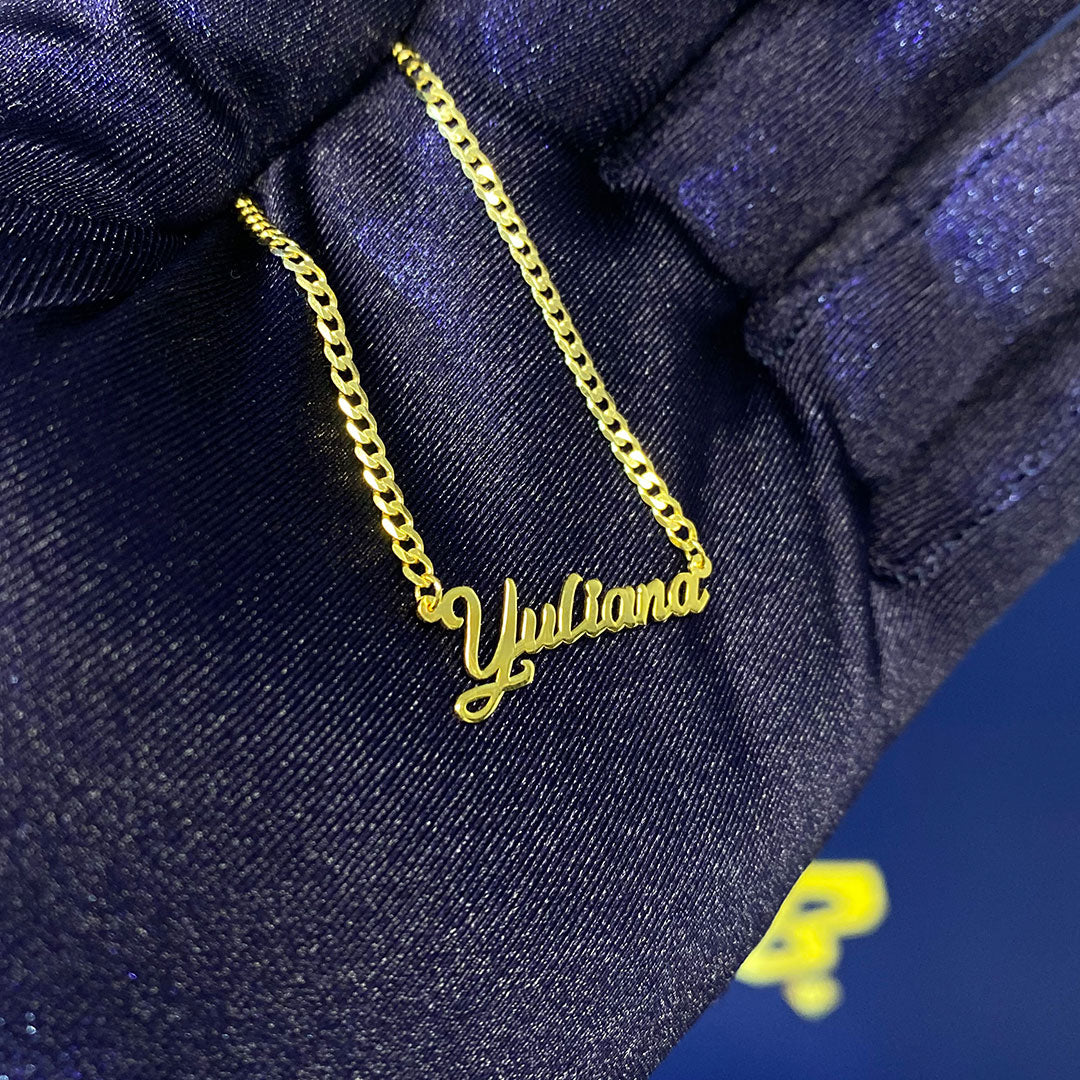 A yellow gold mini pendant with the cursive text 'Yuliana' held in a hand against a blue background.