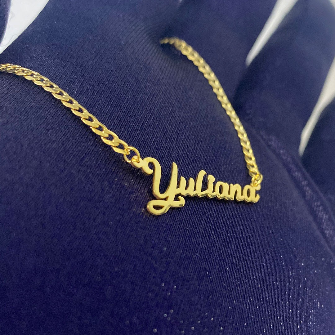 A yellow gold mini pendant with the cursive text 'Yuliana' held in a hand against a blue background. close