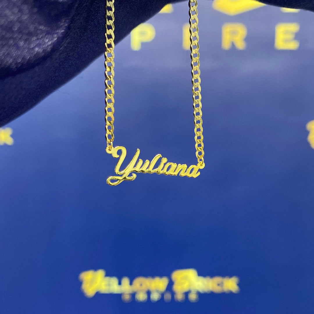 A yellow gold mini pendant with the cursive text 'Yuliana' held in a hand against a blue background.